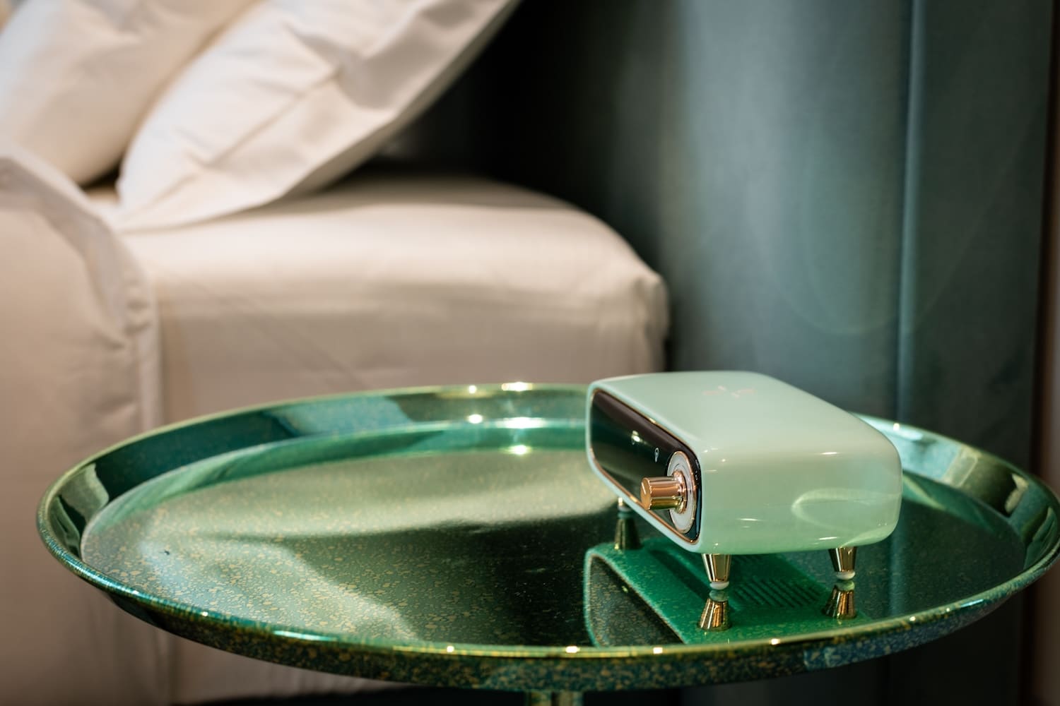 Small retro-style mint green radio with gold knobs on a shiny green round side table next to a bed with white pillows and sheets.