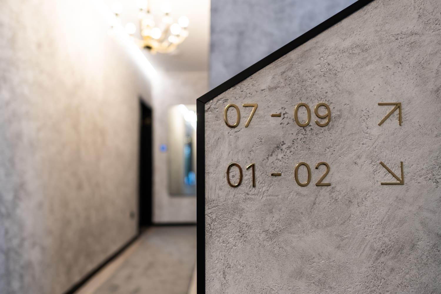 Indoor hallway corner with room number signs 07-09 and 01-02 in gold letters and arrows on textured gray wall.