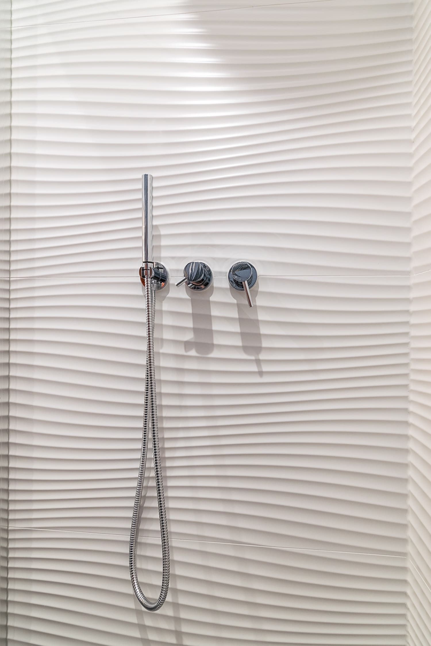 Modern chrome handheld showerhead and two round knobs mounted on white textured wall with horizontal wavy patterns.