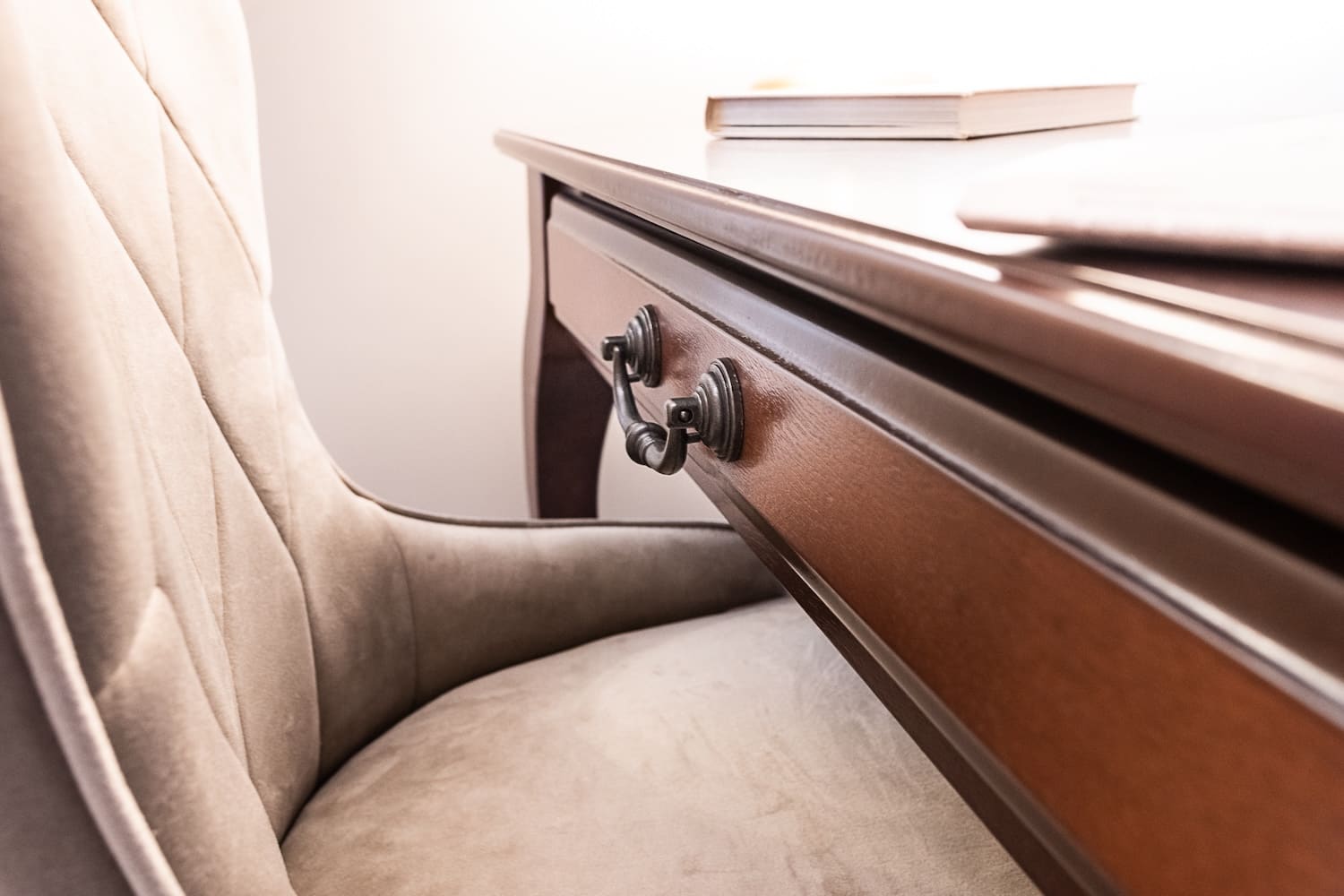 Close-up of a beige upholstered chair next to a wooden desk drawer with metal handles and books on top.