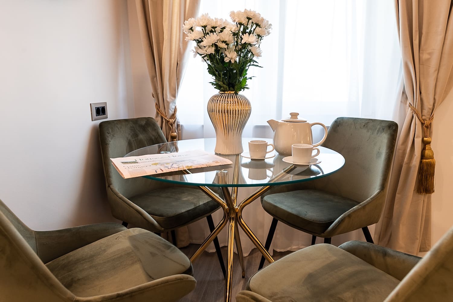 Glass round table with gold legs surrounded by four green velvet chairs, a white vase with white flowers, a teapot, two cups, and a newspaper.