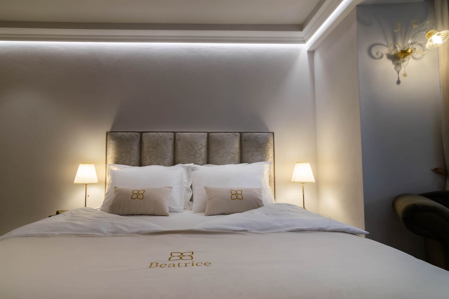 Bed with beige headboard, white pillows, two small beige cushions, and white bedding embroidered with 'Beatrice', lit by two bedside lamps and a wall light.