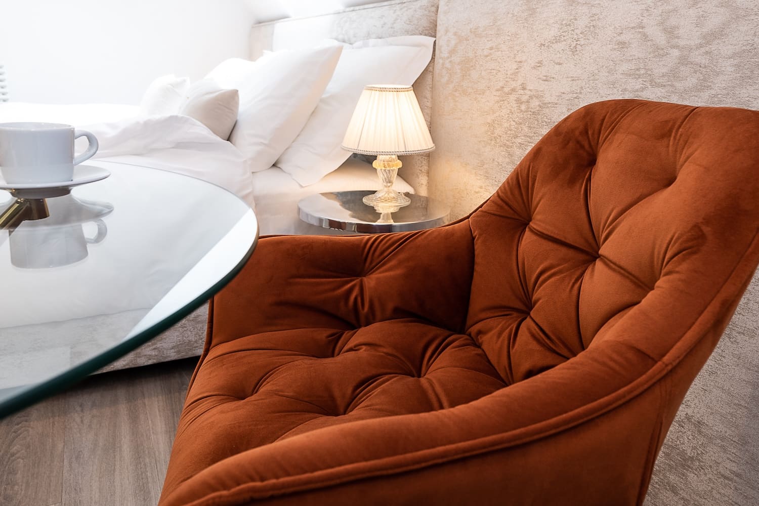 Close-up of a tufted rust-colored armchair next to a glass table with a white cup and saucer, and a bedside lamp on a small round table near a bed with white bedding.