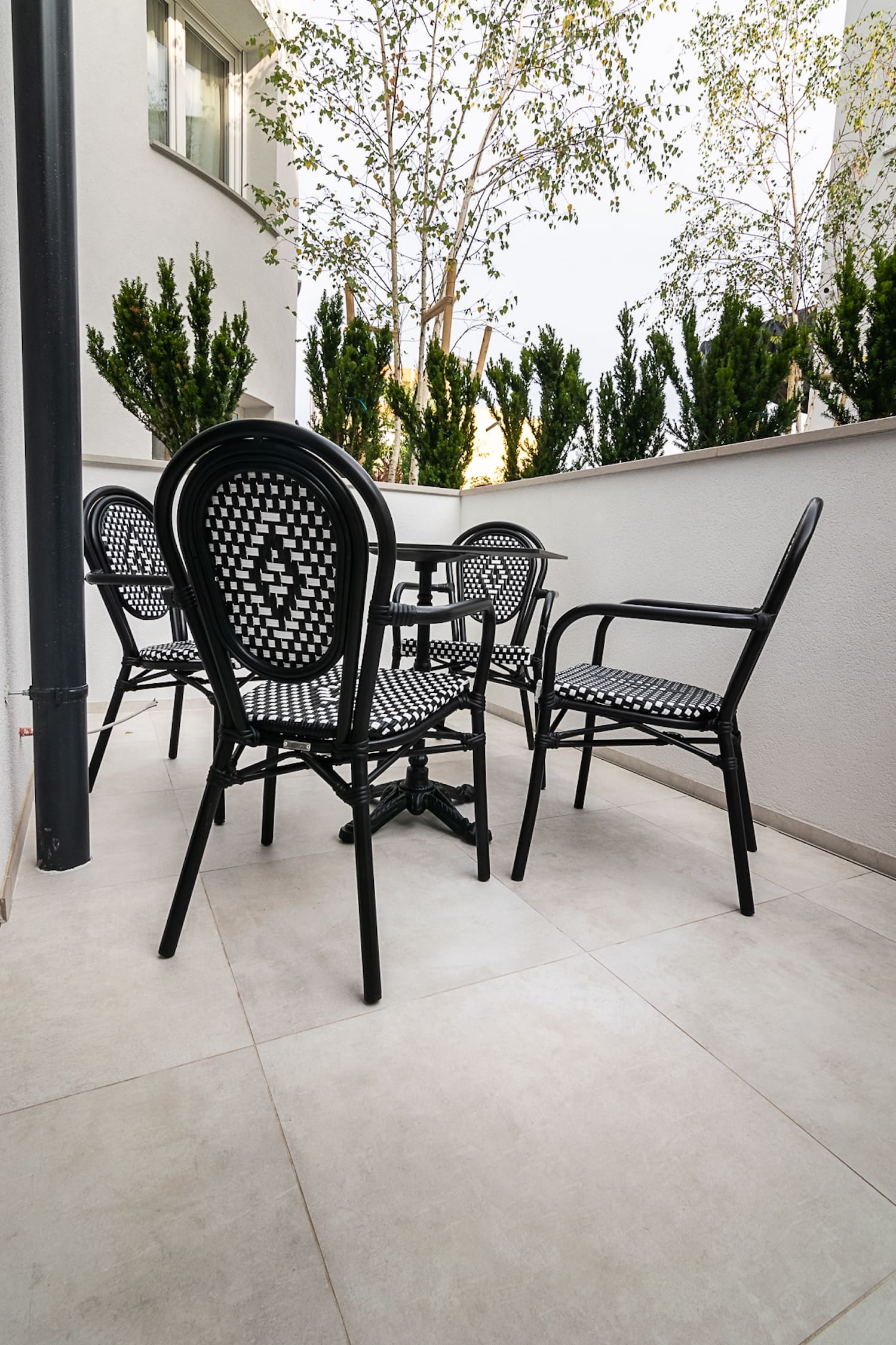 Outdoor patio with four black and white woven chairs around a small black table against a white wall with green shrubs and trees in the background.
