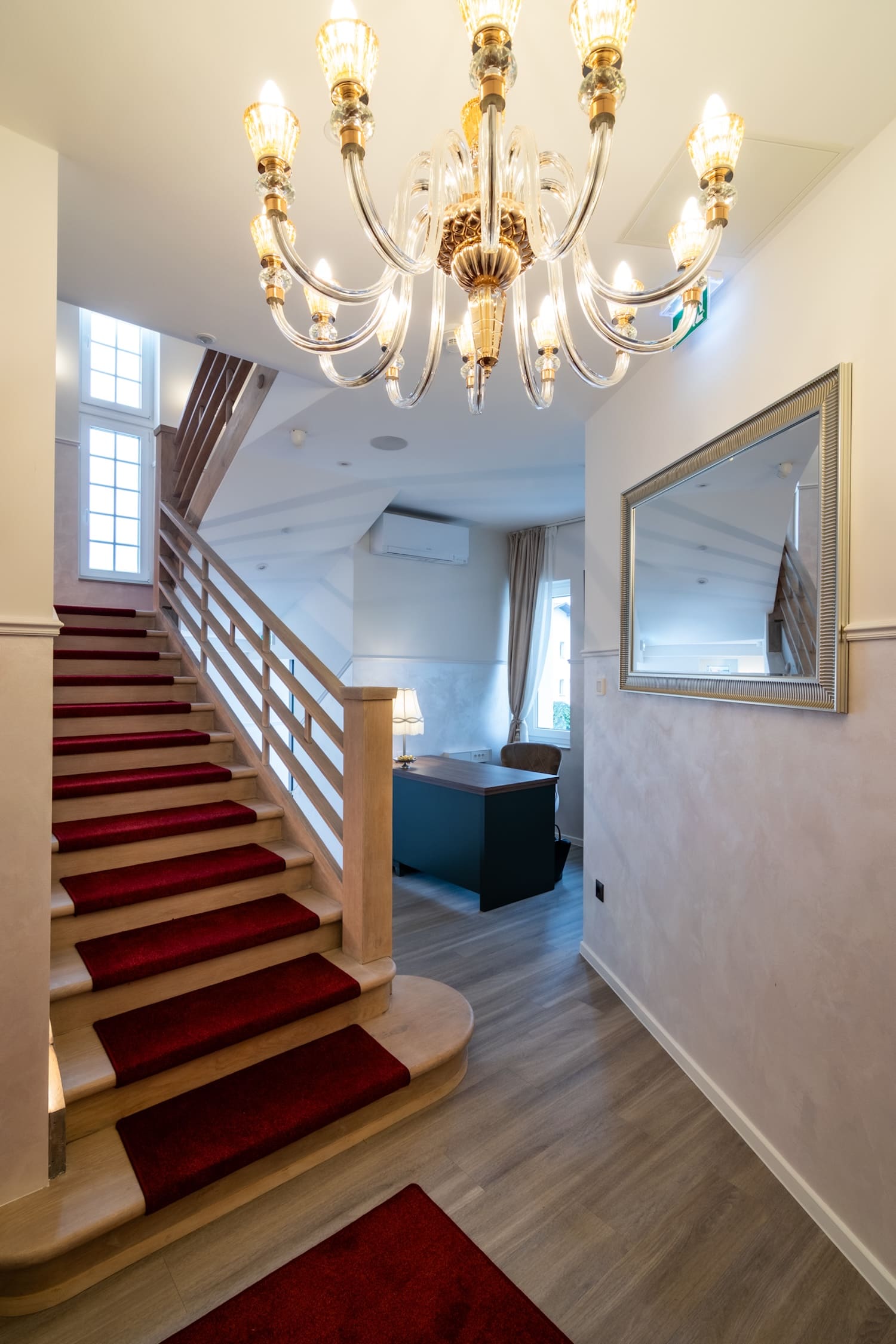 Interior view of a stairway with red carpet runner, wooden banister, chandelier, and a room with desk and window in the background.