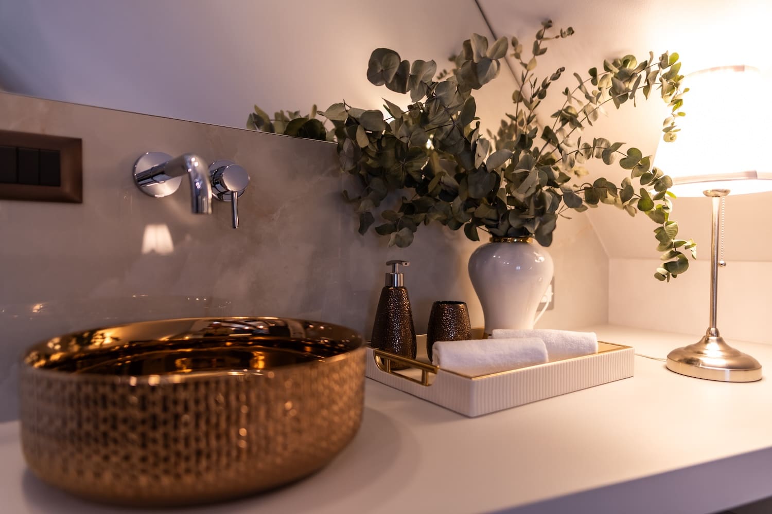 Modern bathroom sink with a gold textured basin, wall-mounted faucet, and decorative tray holding a white vase with eucalyptus, soap dispenser, and towels, next to a table lamp.