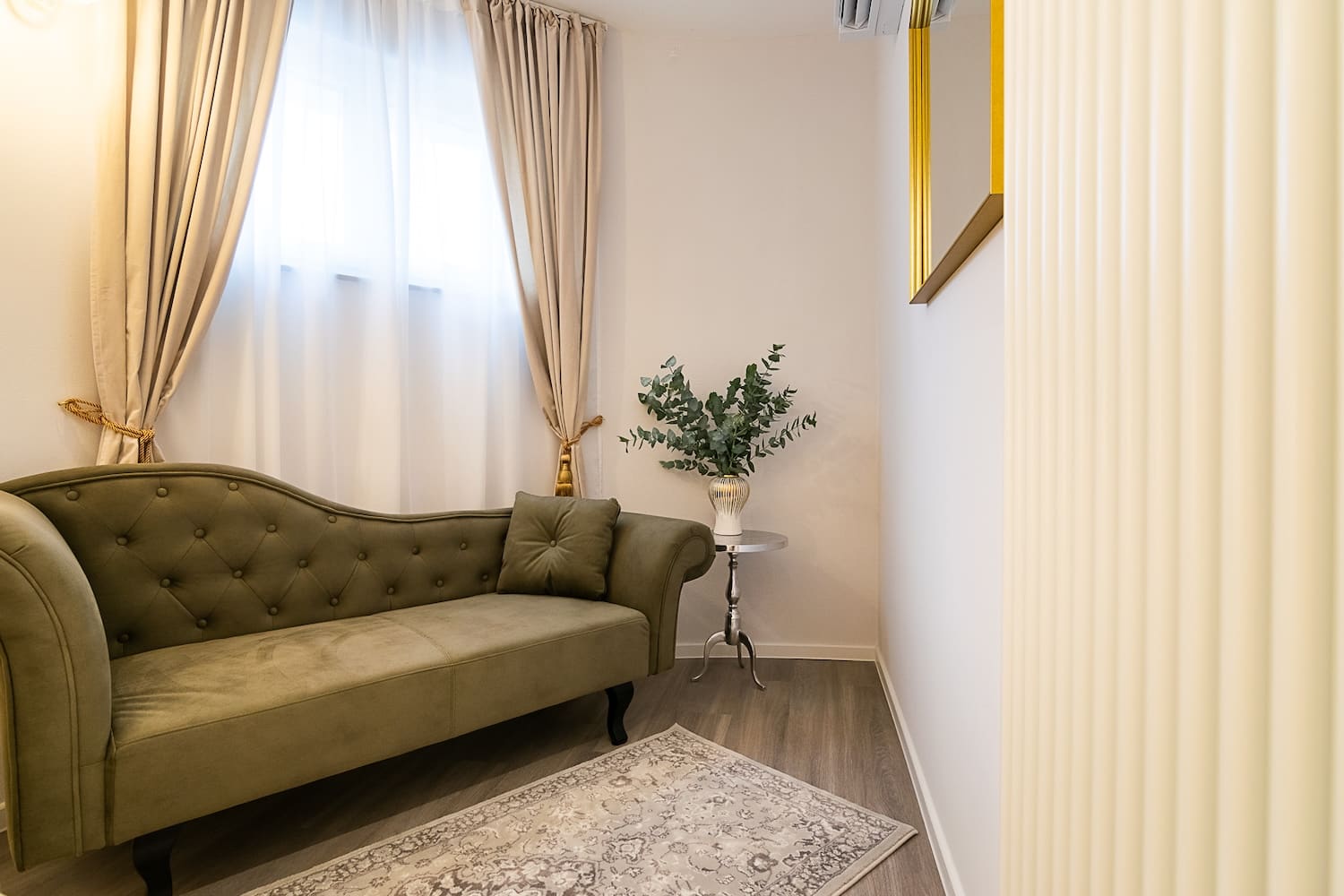 Small cozy room with an olive green tufted sofa, beige curtains, a side table with a vase of greenery, and a patterned rug.