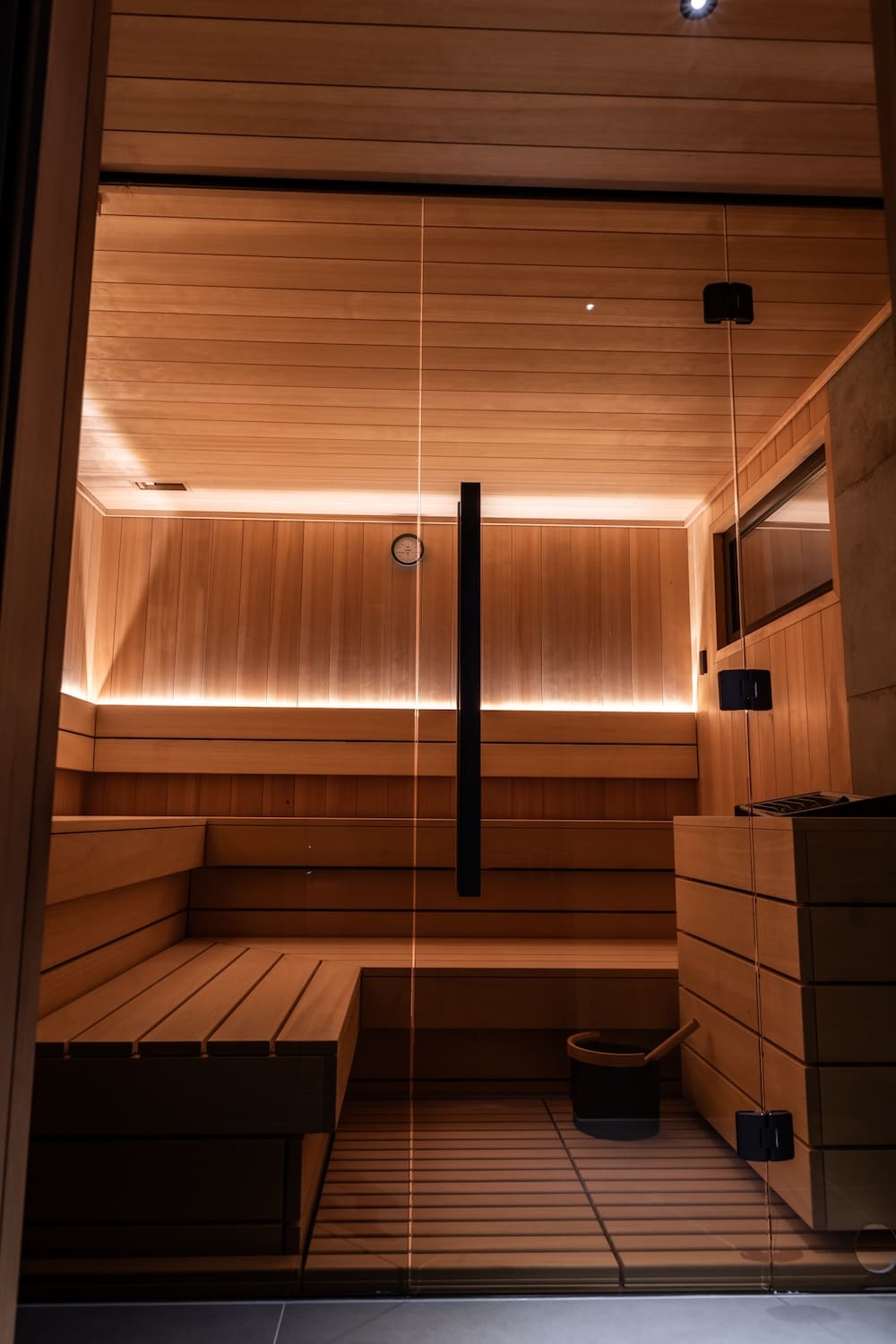 Interior of a modern wooden sauna with built-in benches, glass door with handle, and sauna bucket with ladle.