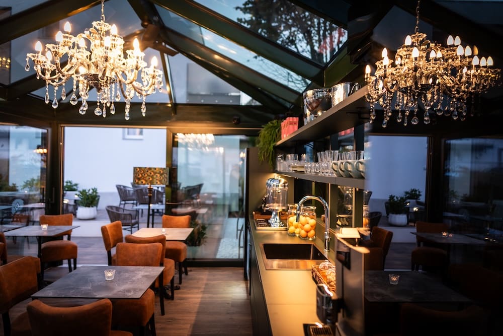 Cozy indoor café with brown chairs, dark tables, gourmet kitchen counter, and two ornate chandeliers.