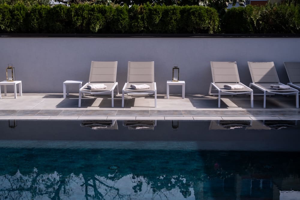 Modern poolside lounge chairs with towels on a tiled deck beside a calm swimming pool reflecting greenery.