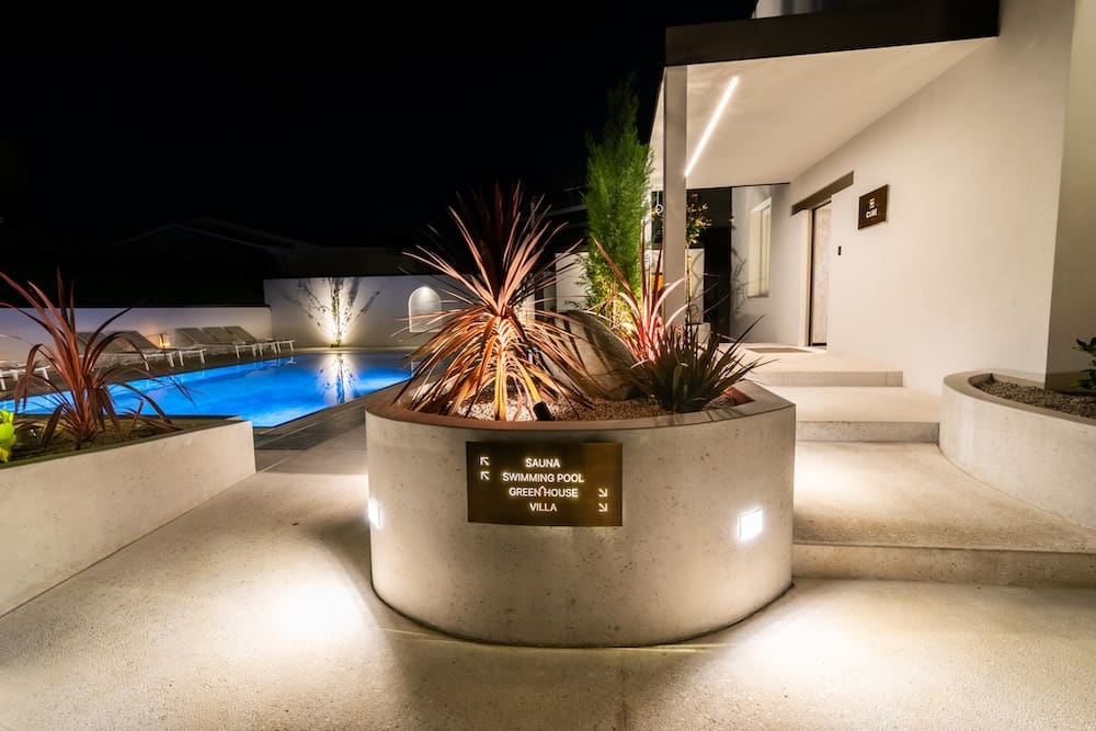 Night view of a modern outdoor area with illuminated swimming pool, concrete planters with decorative plants, and a sign pointing to sauna, swimming pool, greenhouse, and villa.