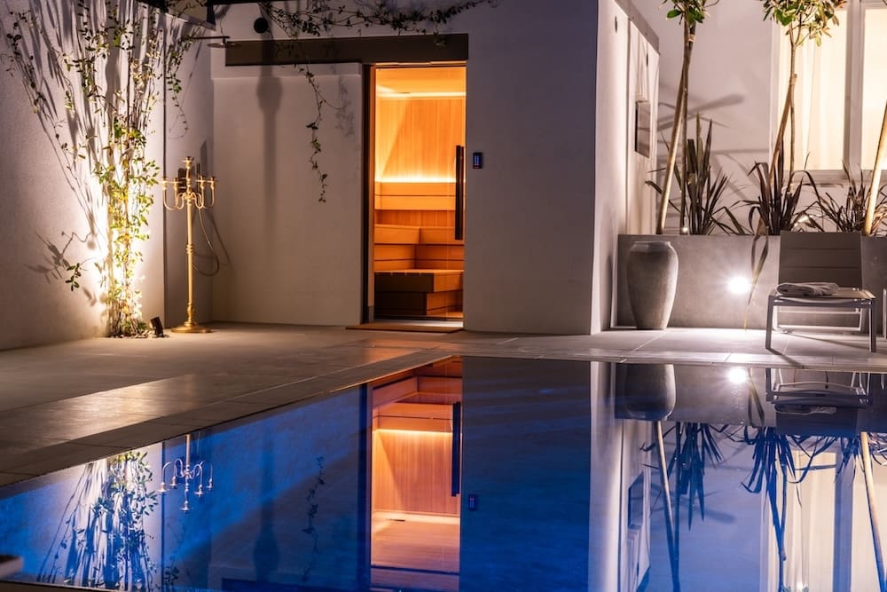 Night view of a modern outdoor pool area with illuminated sauna, plants, and a lounge chair reflecting in the water.