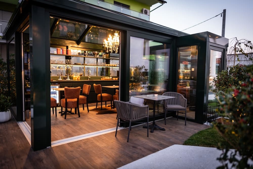 Modern outdoor patio seating area with a glass-enclosed cafe interior featuring brown chairs and warm lighting.