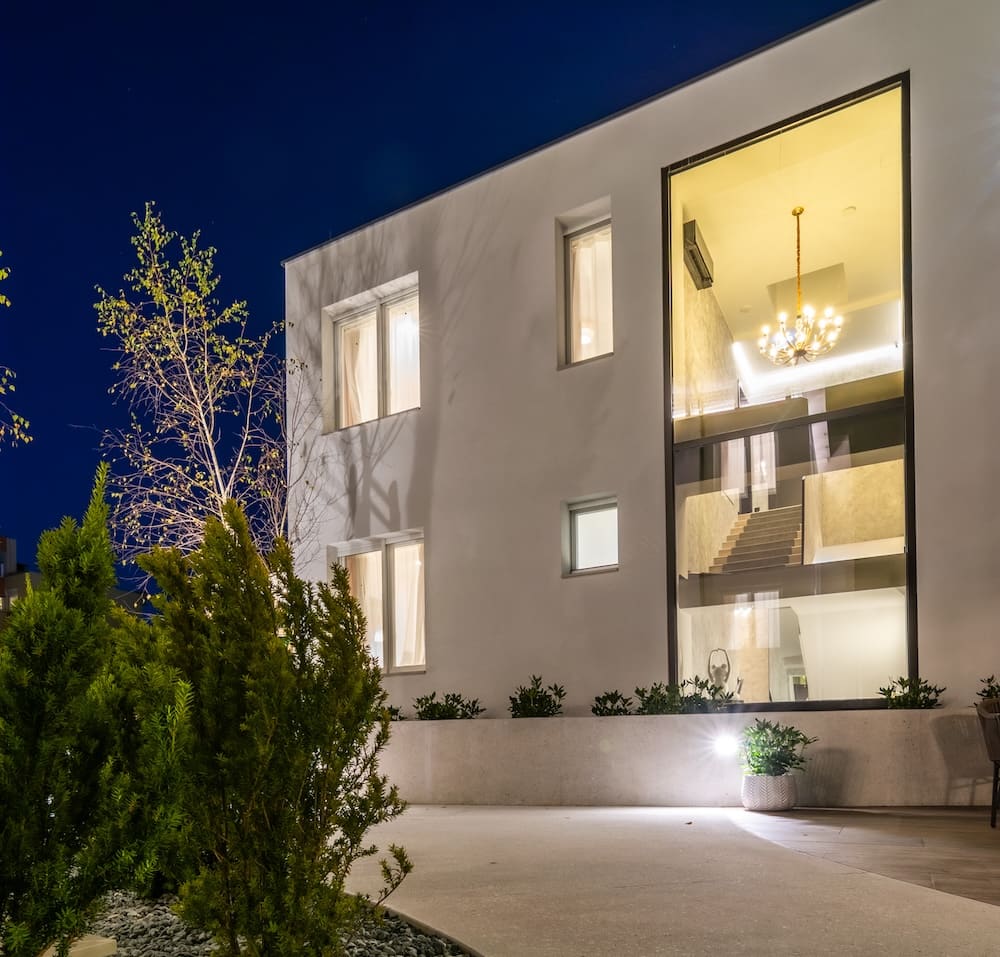 Modern white two-story building with large illuminated window showing staircase and chandelier inside at night.