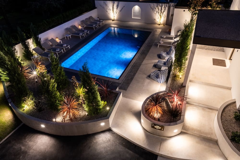 Illuminated modern outdoor swimming pool at night surrounded by lounge chairs, striped bean bags, and landscaped plants.