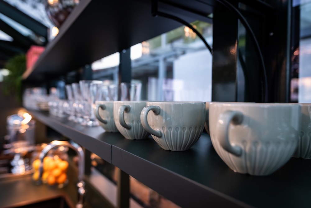 Row of white ceramic mugs and clear glasses neatly arranged on a dark kitchen shelf.