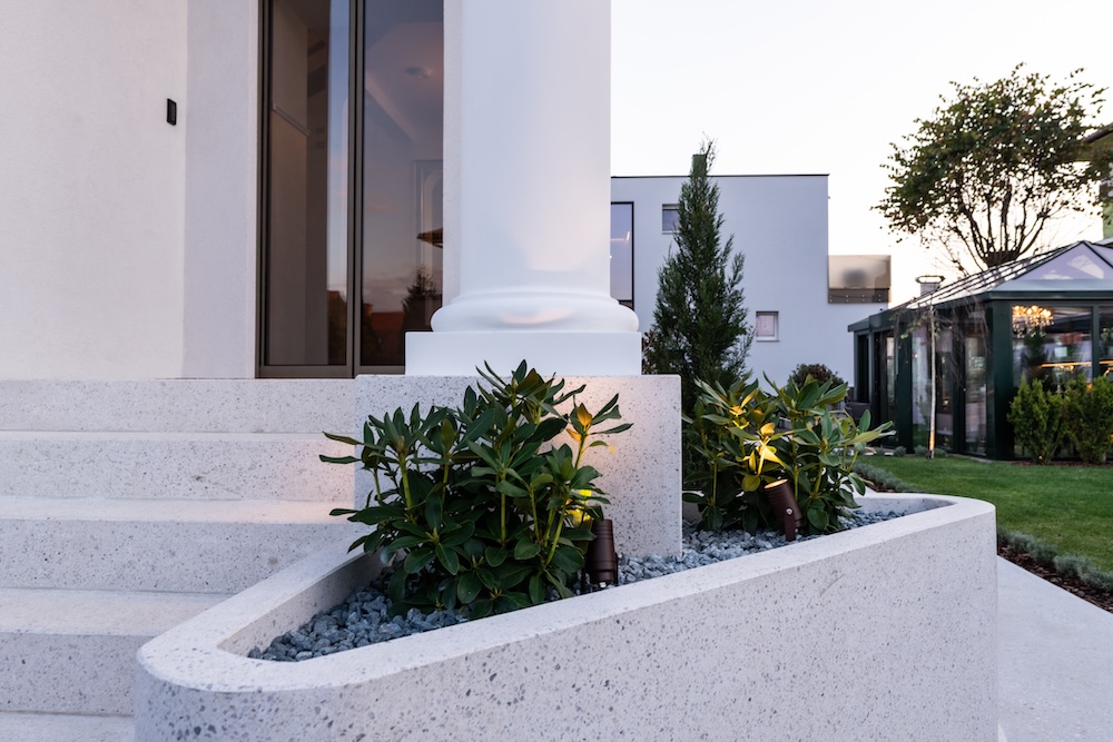 Modern white building entrance with stone steps, large white column, and planted shrubbery illuminated by ground lights.