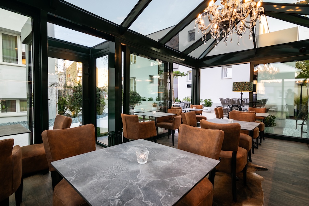 Modern indoor dining area with marble-top tables, brown upholstered chairs, glass walls, and a crystal chandelier.