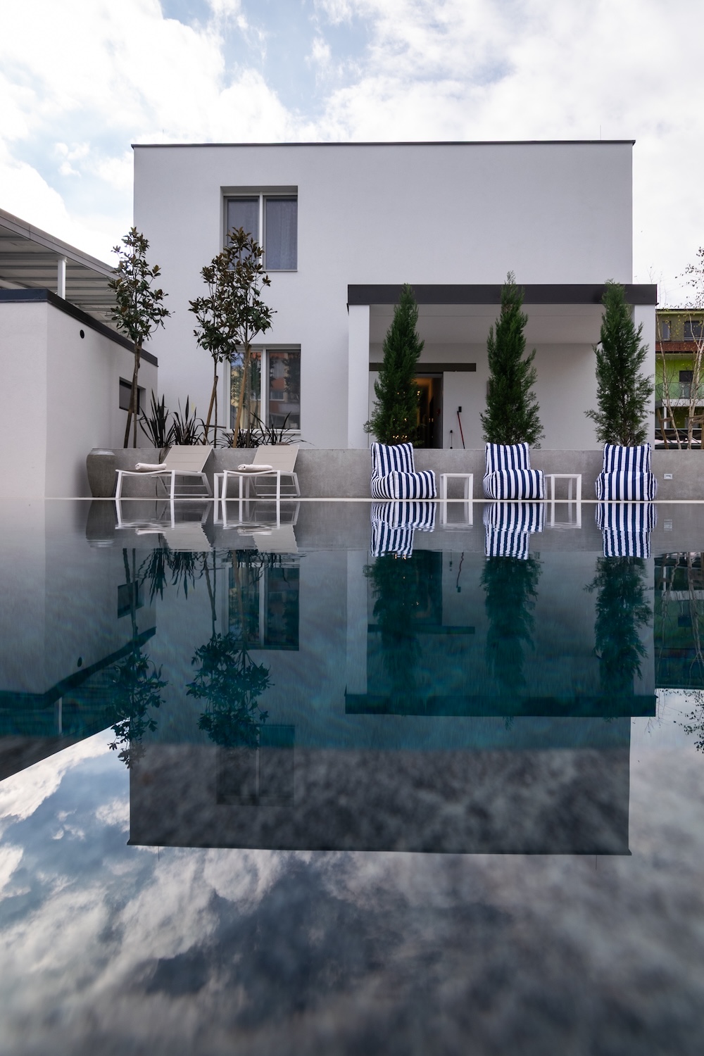 Modern white house with outdoor pool, three striped lounge chairs, two beige chairs, and reflections in the water.
