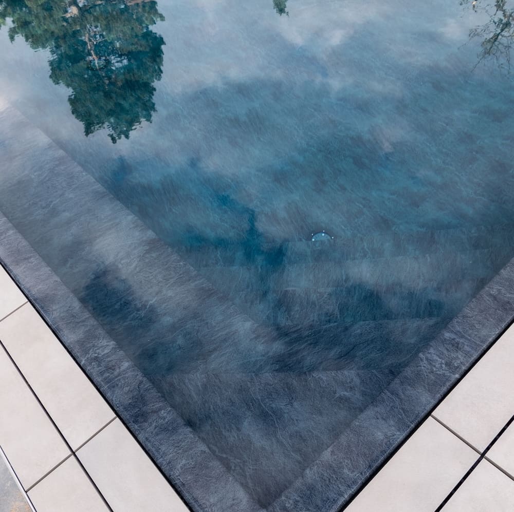 Corner of a tiled swimming pool with clear water reflecting trees and sky.