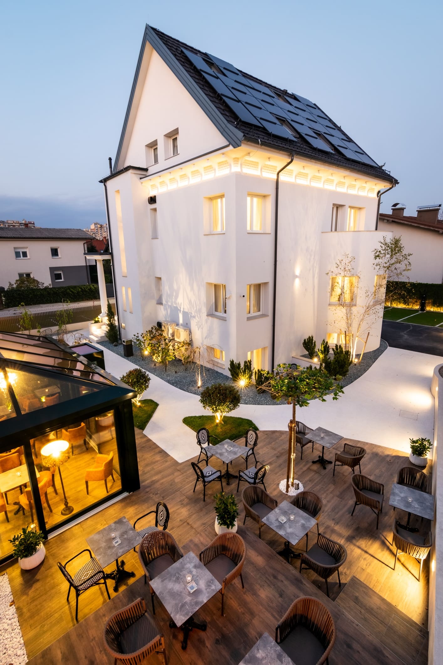 Outdoor patio with tables and chairs beside a modern white house featuring solar panels on the roof at dusk.