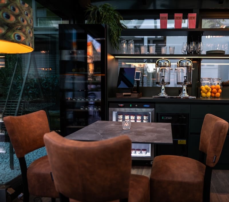 Cozy café seating with four brown chairs around a dark square table, with beverage dispensers, glasses, and fruit on shelves in the background.