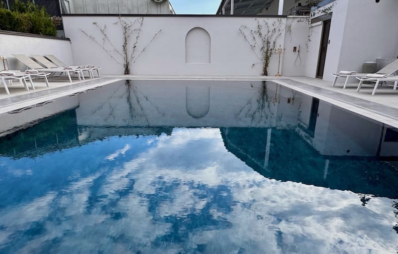Outdoor swimming pool with clear water reflecting a partly cloudy sky, surrounded by white walls and lounge chairs.