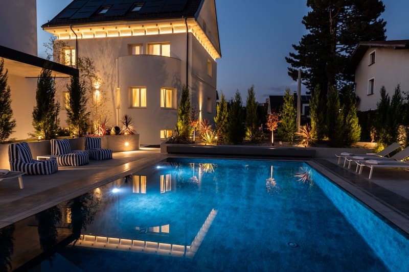 Modern house with warmly lit windows reflected in an illuminated outdoor swimming pool at dusk.