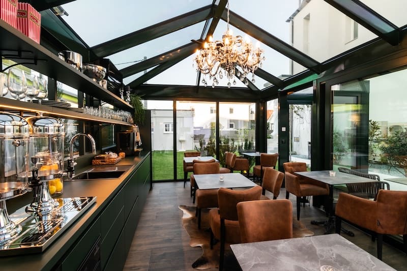Modern glass-walled dining area with brown chairs, marble tables, a chandelier, and a kitchen counter with glass dispensers and shelves.