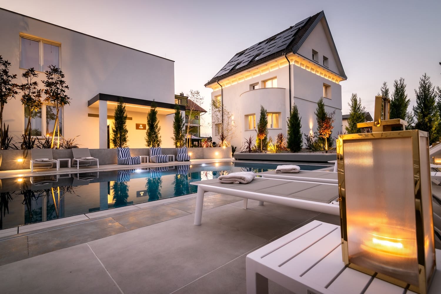 Modern outdoor pool area with white lounge chairs, striped cushions, and illuminated white buildings at dusk.