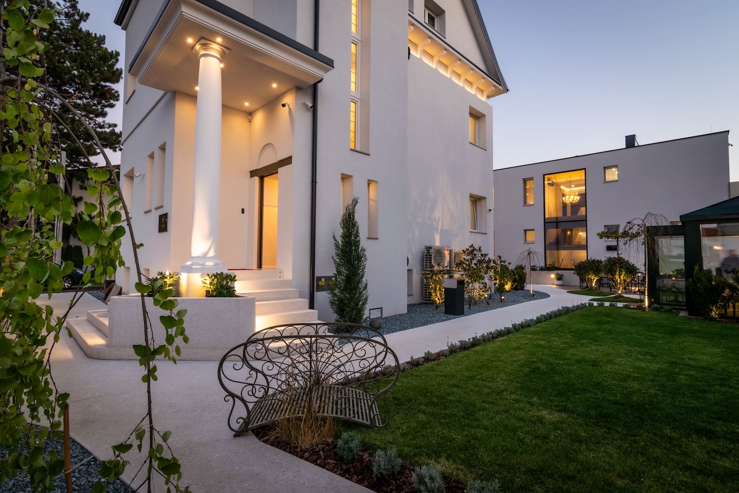 Modern white two-story house with illuminated entrance columns, landscaped garden, and green lawn at dusk.