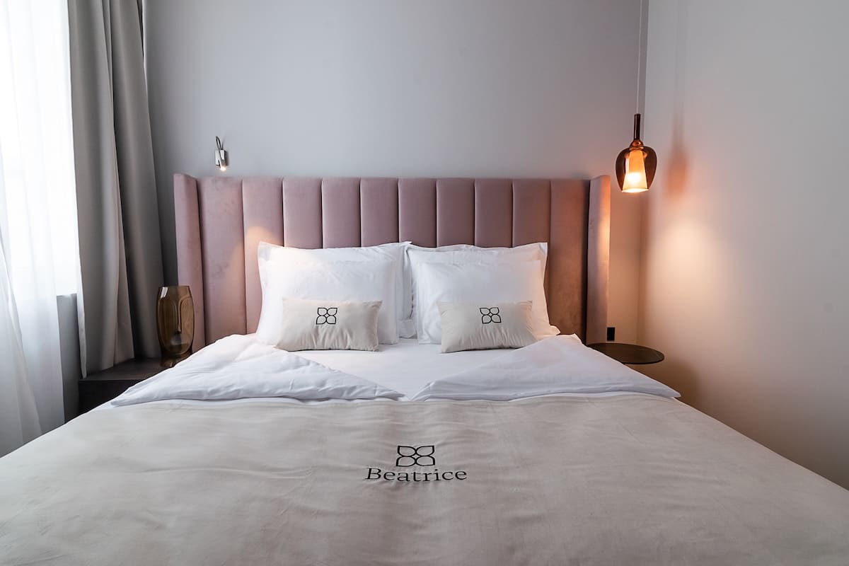Modern bedroom with a large bed featuring white and beige bedding embroidered with 'Beatrice,' a pink upholstered headboard, a hanging lamp on the right, and a sheer curtain on the left.