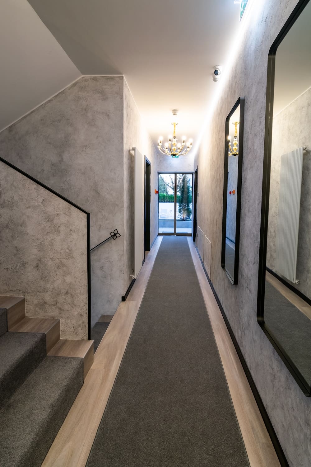 Narrow hallway with gray carpet runner, textured gray walls, staircase on the left, large mirror on the right, and glass door at the end.