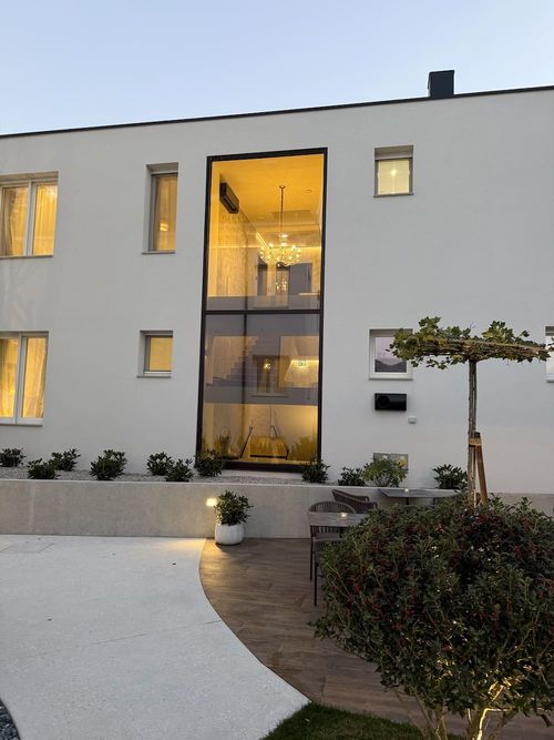 Modern white two-story building with a large window showing an illuminated chandelier inside, small plants along the facade, and an outdoor seating area with chairs and a table under a tree.