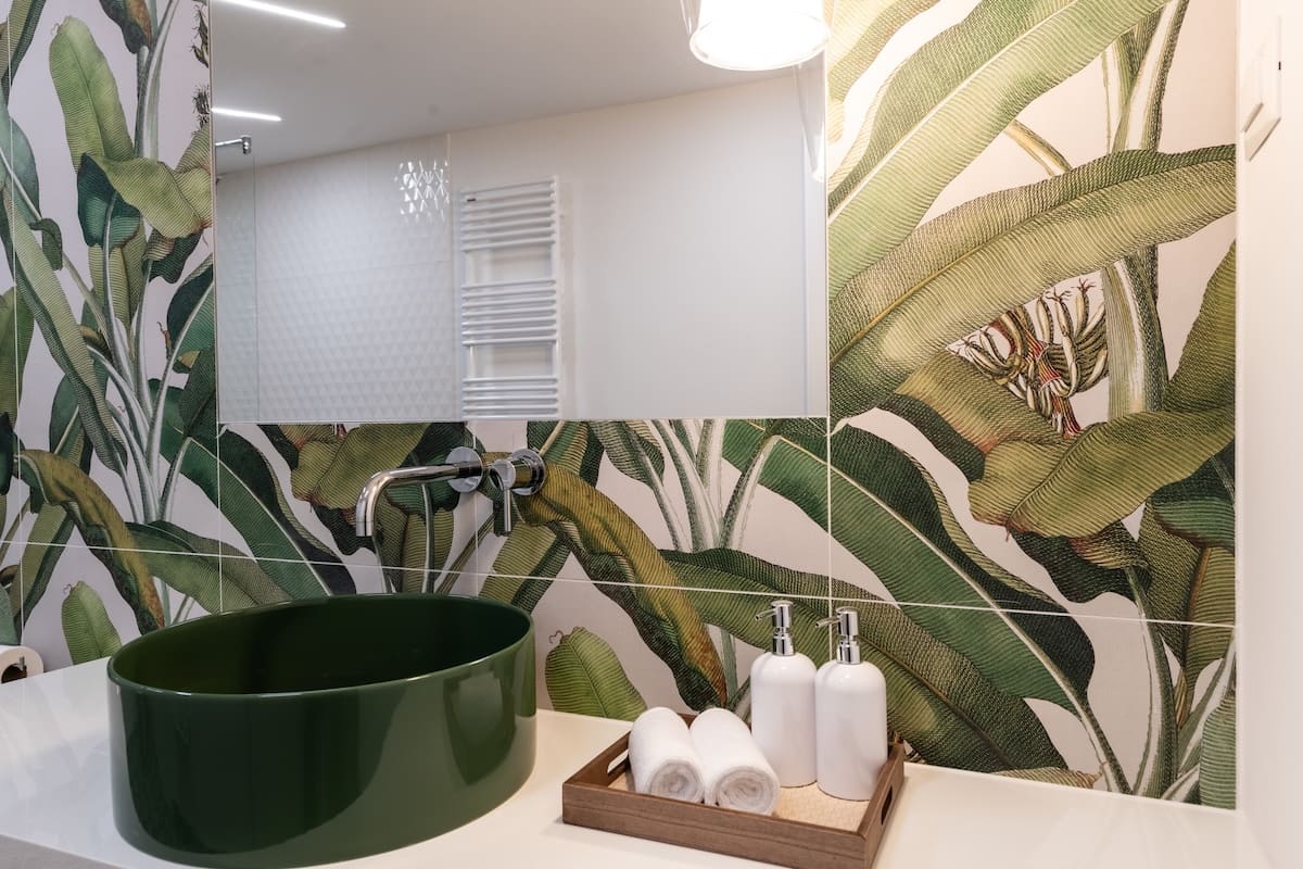 Modern bathroom sink with a dark green oval basin, tropical leaf patterned wall tiles, and a wooden tray holding two white rolled towels and two white pump bottles.
