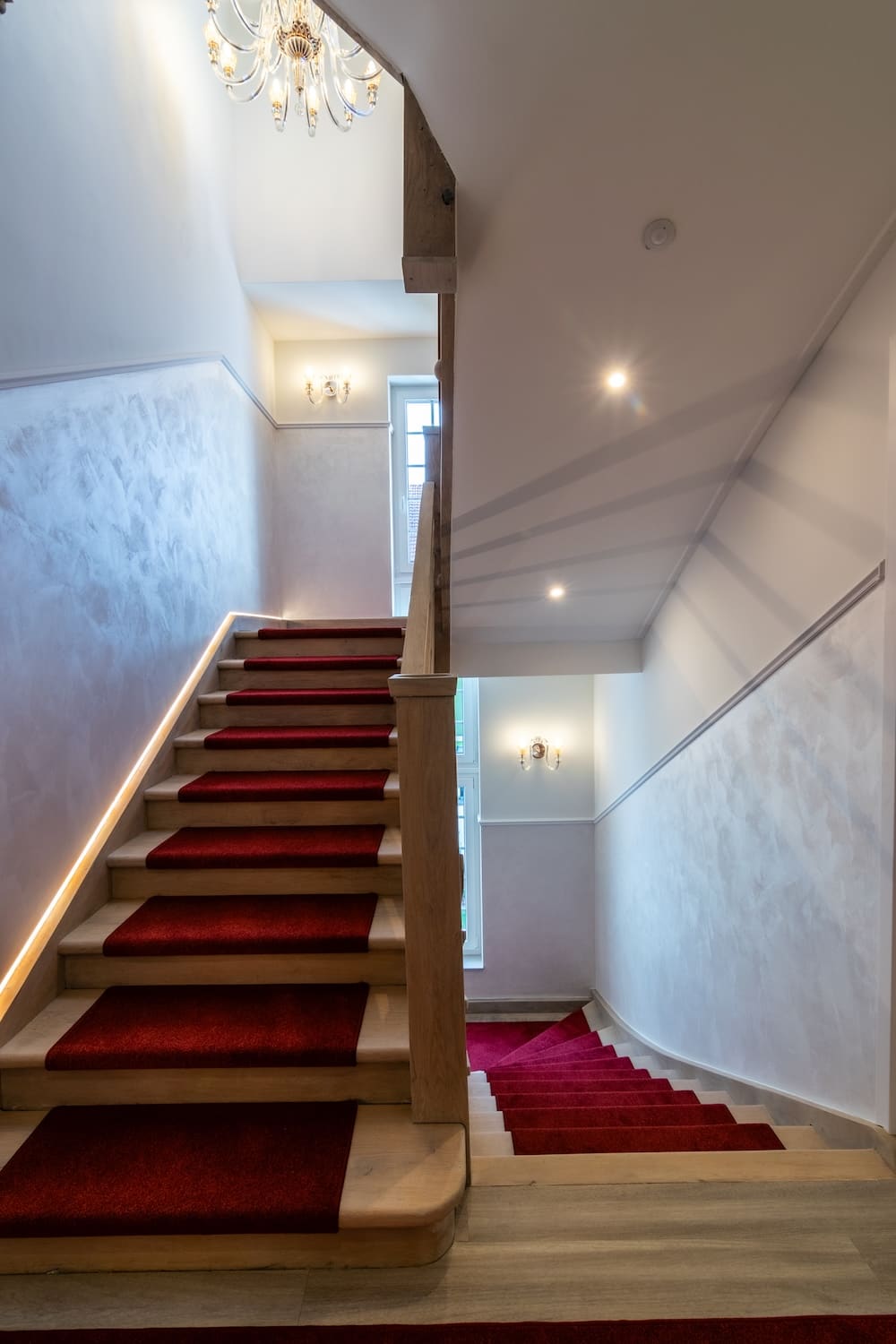 Indoor staircase with wooden steps and red carpet runners, illuminated by wall sconces and a chandelier.