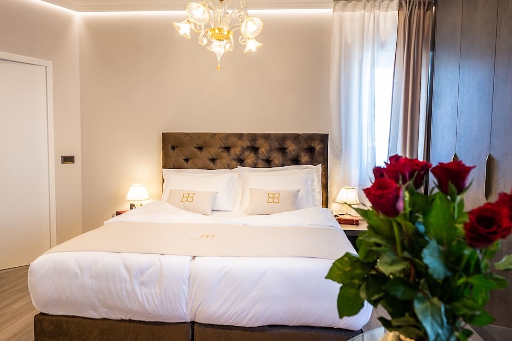 Bright bedroom with a large bed featuring a tufted headboard, white bedding, side tables with lamps, and a bouquet of red roses in the foreground.