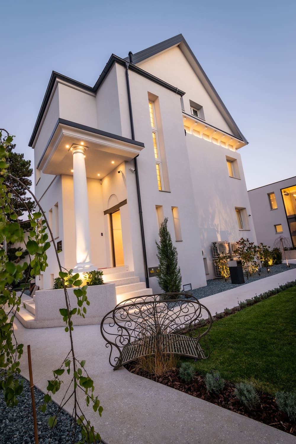 Modern white house with tall columns at entrance, lit warmly at dusk, surrounded by landscaped garden and wrought iron bench.