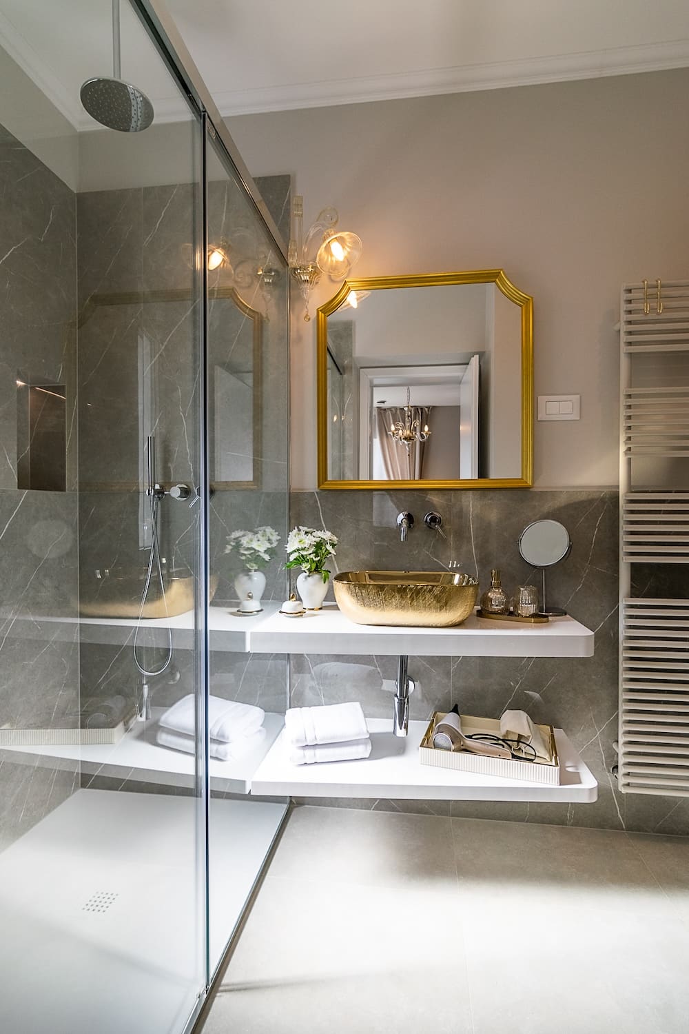 Modern bathroom with glass shower, gold sink and mirror, white shelves holding towels and toiletries.
