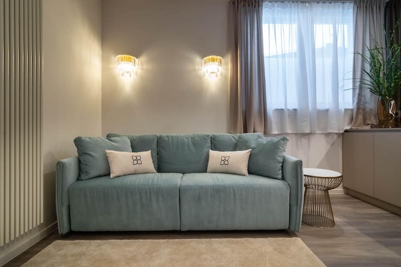 Modern living room with a teal sofa, two decorative pillows, wall sconces, sheer curtains, and a side table with a plant.
