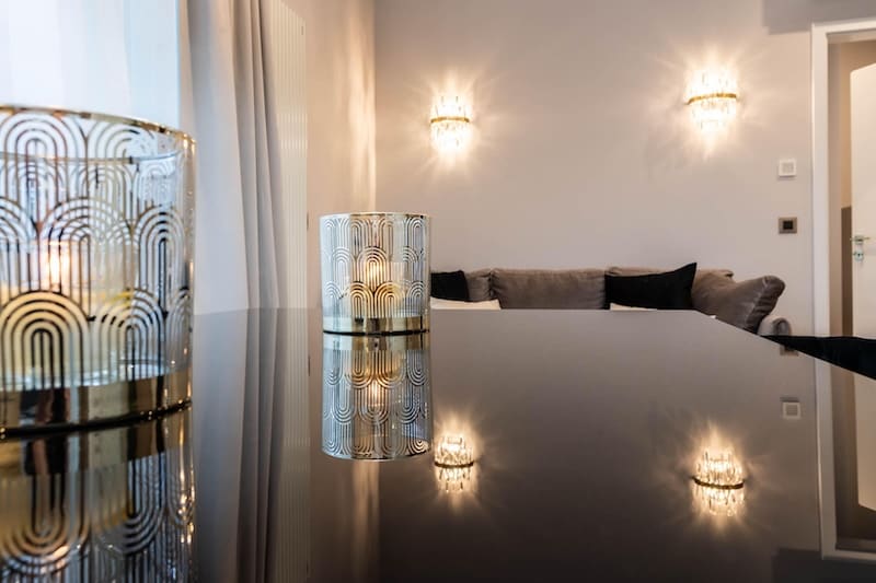 Two decorative candle holders with lit candles on a reflective black surface in a modern living room with a gray sofa and wall sconces.