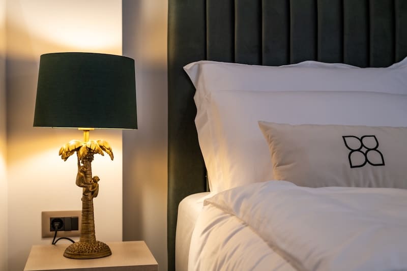 Table lamp with a dark green shade and a palm tree base on a wooden nightstand next to a bed with white linens and a dark green upholstered headboard.