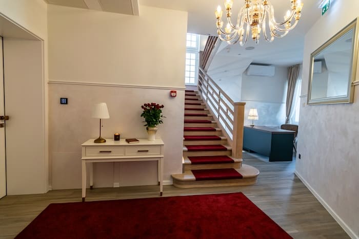 Interior hallway with wooden staircase covered by red carpet runner, white console table with lamp and vase of roses, and chandelier overhead.