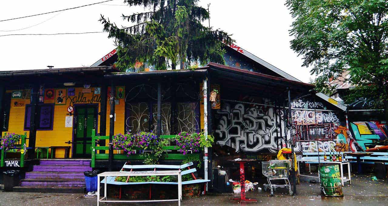 A colorful building with graffiti and flower boxes, featuring a purple staircase, green door, and benches in front under a large tree.