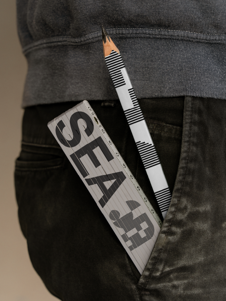 Mètre pliant gris au logo SEA Architecture et crayon rayé noir et blanc glissés dans une poche de jean