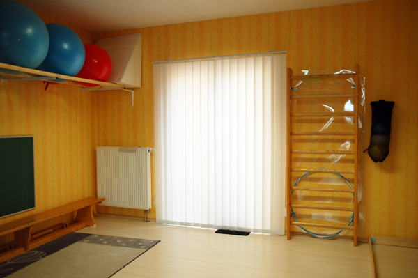 Turnhalle mit Gymnastikbällen auf einem Regal, Wandstab und Vorhängen vor einem Fenster.