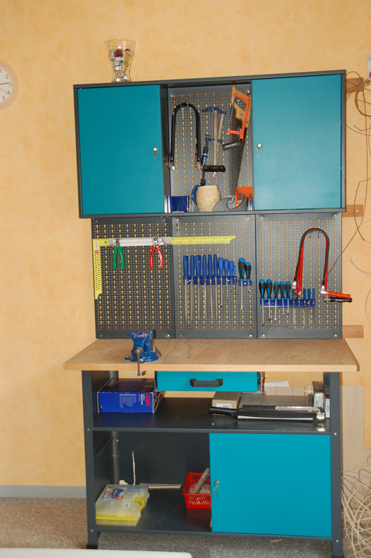 Werkbank mit Holzplatte, blauen Schränken, Schraubendrehern, Zangen, einer Bügelsäge und anderen Werkzeugen an der Wand.