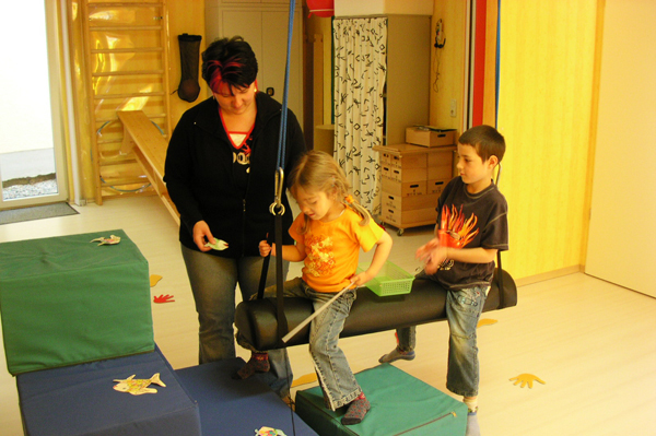 Eine Frau betreut zwei Kinder beim Klettern auf weichen Matten und einer Schaukelsitz in einem hellen Raum.