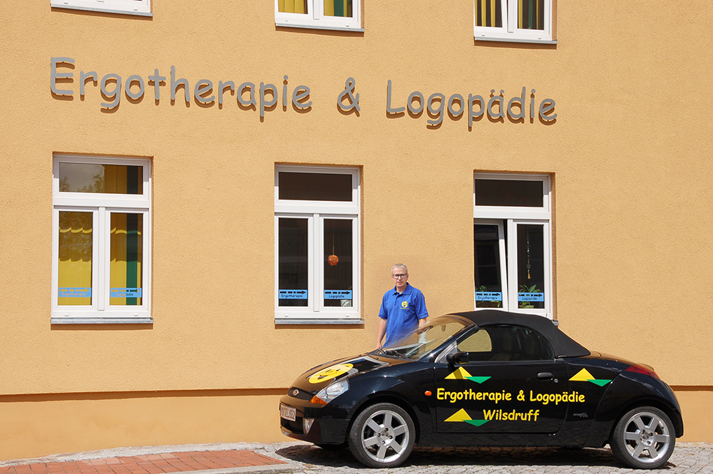 Ein Mann in blauem Hemd steht vor einem gelben Gebäude mit der Aufschrift ‚Ergotherapie & Logopädie‘ und einem schwarzen Auto mit der gleichen Beschriftung.