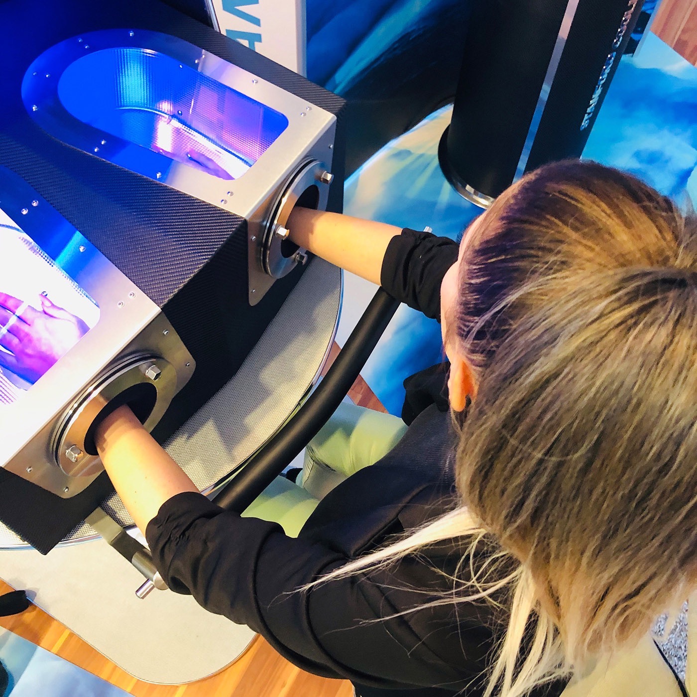 Eine Frau mit einem Pferdeschwanz steckt ihre Hände in ein Gerät mit blauer Beleuchtung, möglicherweise zur Haut- oder Handanalyse.